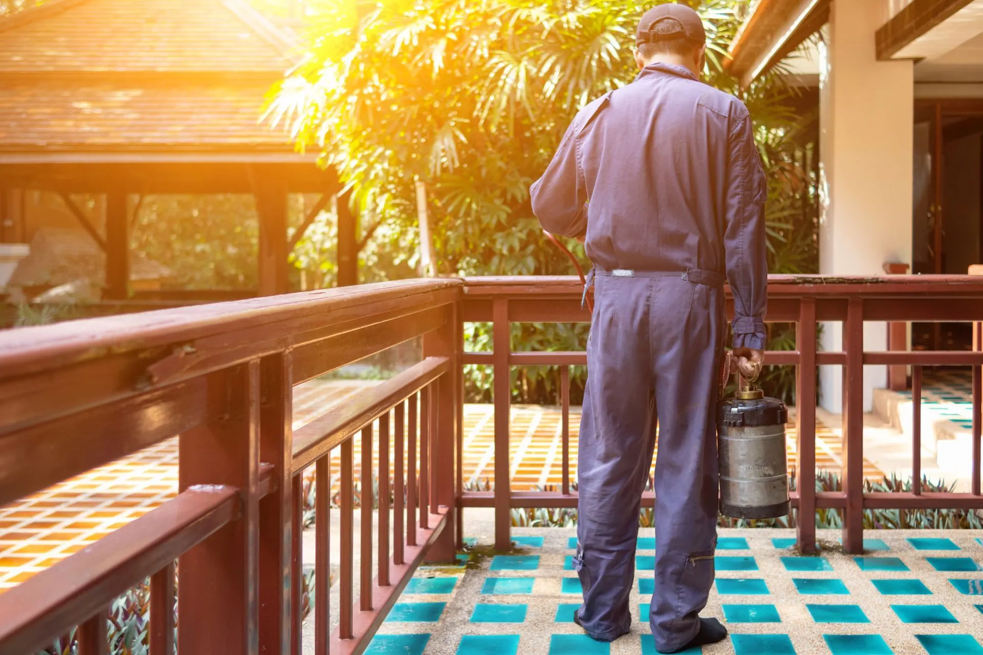Pest control technician in navy coveralls carrying a pressure sprayer along a garden terrace