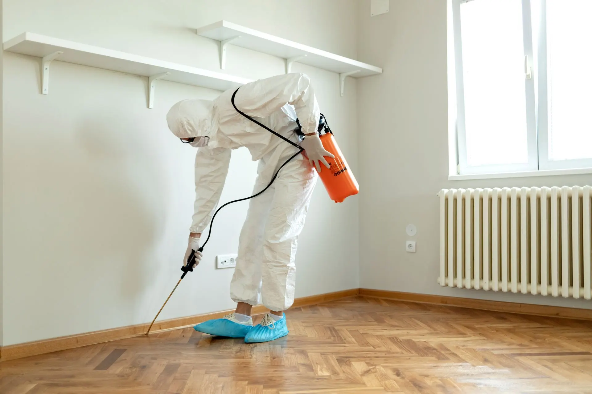 Technician in coveralls spraying termite treatment along a room baseboard
